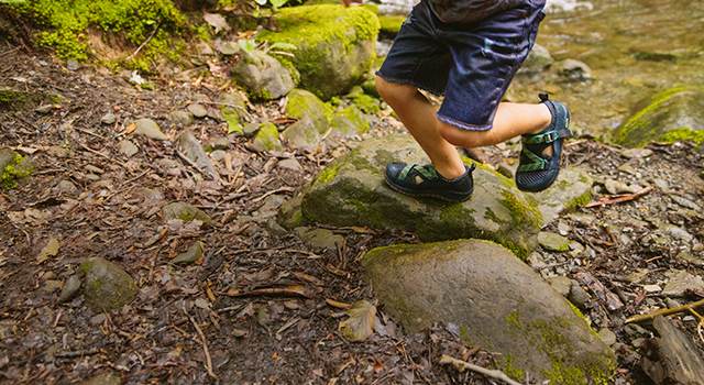 Kid runs on top of rocks in the woods while wearing Odyssey sandals.