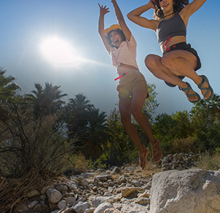 Two females jumping from rocks in the midday sun, with big smiles on their faces and Chaco sandals on their feet.