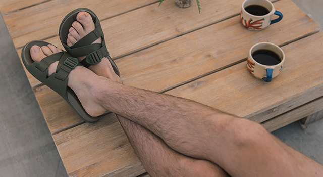A man sits with legs crossed and feet up on table with cups of coffee and his Chillos slide sandals.